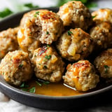 A close-up view of golden-brown meatballs garnished with herbs, some containing melted cheese, served in a dark bowl with a light sauce.