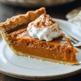 A slice of pumpkin pie topped with whipped cream and a dusting of cinnamon, served on a beige plate.