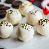 A close-up of white chocolate-covered cake balls adorned with colorful sprinkles, placed on parchment paper next to chocolate cookies.