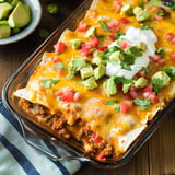 A baked dish of cheese-covered enchiladas topped with diced tomatoes, avocado, cilantro, and sour cream, served on a wooden table alongside sliced jalapeño.