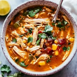 A bowl of chicken soup featuring shredded chicken, corn, diced tomatoes, and cilantro, garnished with pieces of green pepper and served with a slice of lime on the side.