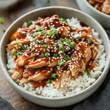 A bowl of shredded chicken topped with green onions and sesame seeds, served over a bed of white rice and drizzled with a savory sauce.