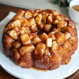 A round bundt cake topped with caramelized apples and a glossy brown sauce sits on a white plate.