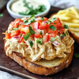 A slice of toasted bread topped with shredded chicken, diced tomatoes, cilantro, and red onions, served with a bowl of sauce and French fries in the background.