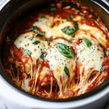 A close-up view of chicken topped with melted mozzarella cheese and basil leaves, simmering in a rich tomato sauce inside a slow cooker.