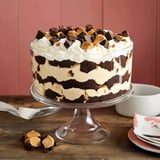 A layered chocolate and vanilla dessert topped with whipped cream and chocolate pieces is displayed in a glass trifle dish.