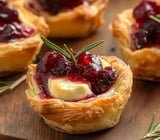 A close-up of a pastry cup filled with creamy cheese and topped with red fruit preserves, garnished with a sprig of rosemary.