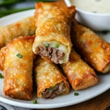 A plate of golden-brown, crispy egg rolls filled with beef and green onions, accompanied by a small bowl of dipping sauce.