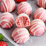 A close-up of pink cake balls with white drizzle, one with a bite taken out, revealing a soft, pink interior.