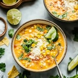 A bowl of corn soup garnished with cilantro, cheese, and lime wedges, accompanied by tortilla chips and salt in the background.