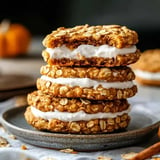 A stack of oatmeal cookies filled with white cream, with one cookie bitten into, placed on a gray plate.