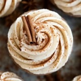 A close-up of a frosted cupcake topped with two cinnamon sticks and a dusting of ground cinnamon.