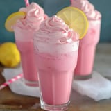 Three tall glasses filled with pink frothy drinks topped with whipped cream and garnished with lemon slices, accompanied by a lemon and a paper straw.