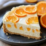 A slice of creamy orange cheesecake topped with fresh orange slices on a black plate.