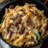 A close-up view of a bowl of creamy pasta with penne, pieces of beef, and green vegetables, garnished with black pepper and cheese.