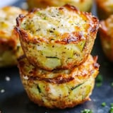 Two crispy zucchini muffins stacked on top of each other, with a golden-brown crust and flecks of green herbs, on a dark surface.