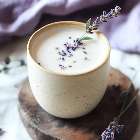 A cup of lavender-infused beverage topped with dried lavender flowers sits on a wooden round surface, surrounded by lavender sprigs and a soft purple fabric in the background.