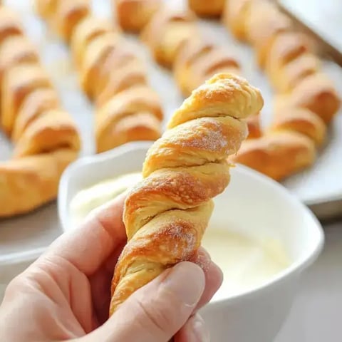 A hand holds a twisted pastry over a bowl of creamy dip, with more twisted pastries in the background.