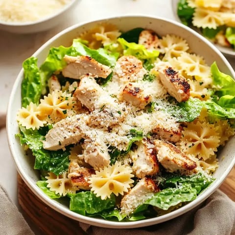 A bowl of salad featuring grilled chicken, romaine lettuce, and bowtie pasta, topped with grated cheese and herbs.