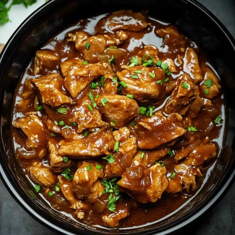 A bowl of tender, diced chicken pieces coated in a rich brown sauce and garnished with chopped green herbs.