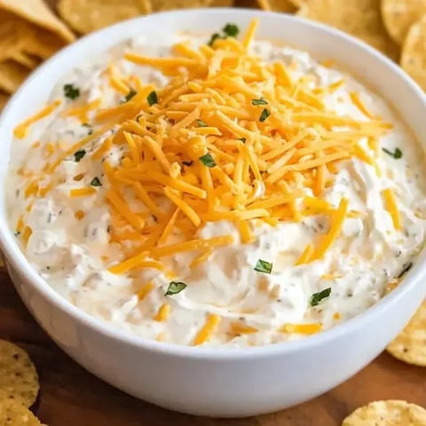 A bowl of creamy dip topped with shredded cheddar cheese and garnished with herbs, surrounded by tortilla chips.